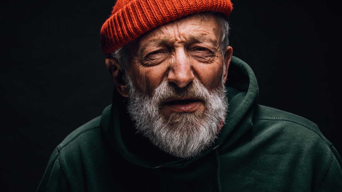 Old homeless man dressed in worn but clean outdoor clothes and hat looking at camera with screwed up eyes and interrogative expression having sight.