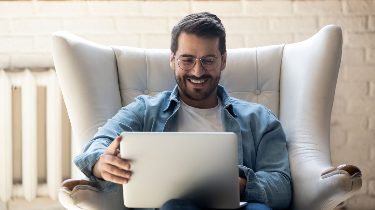 Smiling young man wearing glasses using laptop, sitting in cozy armchair, happy male looking at computer screen, chatting in social network or shopping online, playing game, working at home.