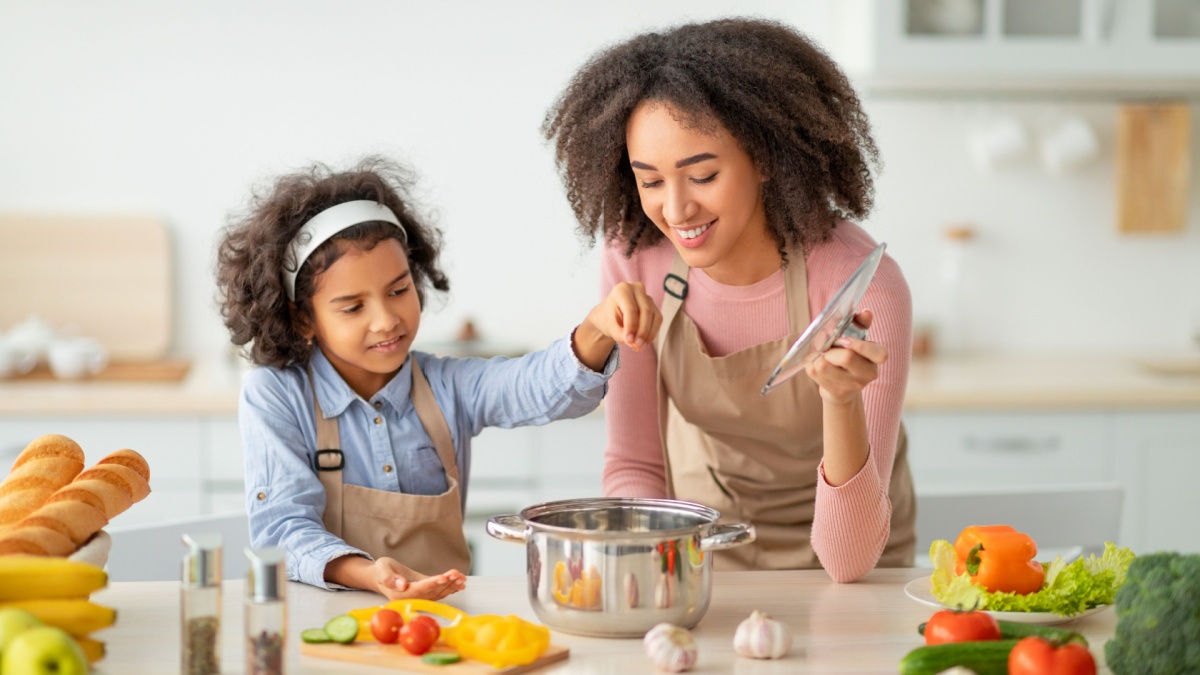 Bonding Concept. Cheerful african american mother helping her daughter to cook, teaching little girl how to prepare healthy vegetable soup, salting water in pot, cooking together at home.