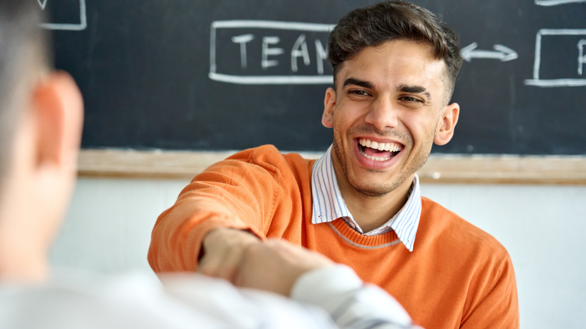 Young happy laughing Indian latin Hispanic creative startup coworker student giving fist bump to classmate partner committing successful project result in office classroom at desk near blackboard.